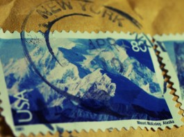Primer plano de dos sellos postales de EE. UU. que muestran el monte McKinley en Alaska, parcialmente oscurecidos por un matasellos de Nueva York, resaltando íconos perdurables incluso en la era del comercio electrónico.