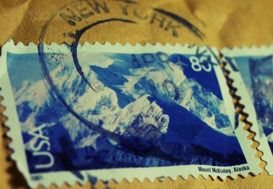 Primer plano de dos sellos postales de EE. UU. que muestran el monte McKinley en Alaska, parcialmente oscurecidos por un matasellos de Nueva York, resaltando íconos perdurables incluso en la era del comercio electrónico.