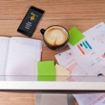 Escritorio de oficina con una agenda abierta, un teléfono inteligente, café, gráficos coloridos en papel, notas adhesivas y un portalápices.