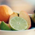 Un recipiente con frutas variadas que incluye un plátano, rodajas de lima, una manzana y partes de una naranja.