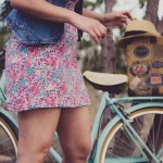 Una persona con un vestido floral y un chaleco vaquero está cerca de una bicicleta azul vintage con una maleta decorada en la parte trasera.