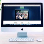 Un iMac sobre un escritorio muestra una ventana del navegador con la imagen de un grupo de personas. A la izquierda hay una planta en maceta con flores amarillas.