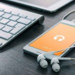 Un teléfono inteligente con auriculares que muestra una aplicación de música se encuentra junto a un teclado y una tableta sobre un escritorio de madera.