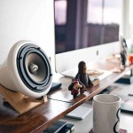 Un altavoz blanco sobre un escritorio de madera con un monitor de computadora y pequeñas figuras cerca, junto a una taza blanca.