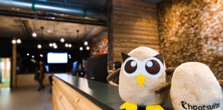 Un búho de peluche se posa sobre un mostrador de recepción de madera, contra una pared de ladrillos difuminados y un techo de estilo industrial, lo que agrega encanto a este fondo con temática de empresas, ideal para exhibir en redes sociales.