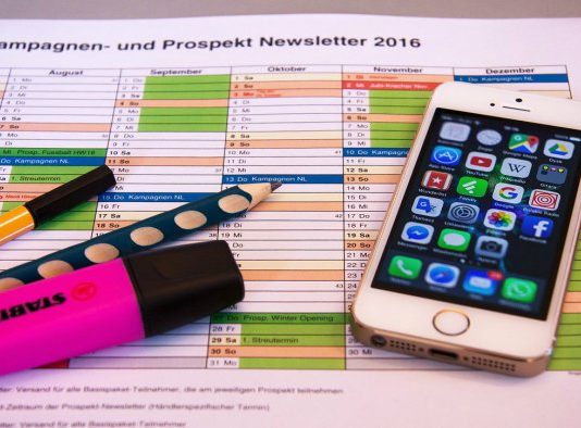 Un calendario con secciones de colores, un teléfono inteligente que muestra aplicaciones de marketing digital, un resaltador rosa, un marcador negro y un bolígrafo con lunares azules se encuentran sobre el escritorio de una empresa innovadora.