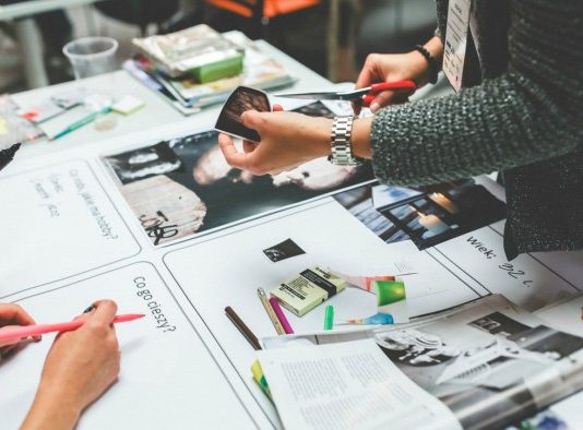 Personas colaborando en una mesa llena de papeles, fotos, marcadores y revistas, intercambiando ideas para un nuevo logotipo de empresa que se destaque de la competencia.