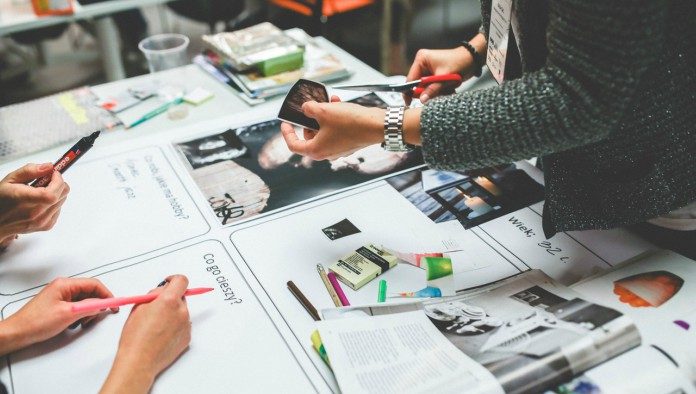 Personas colaborando en una mesa llena de papeles, fotos, marcadores y revistas, intercambiando ideas para un nuevo logotipo de empresa que se destaque de la competencia.