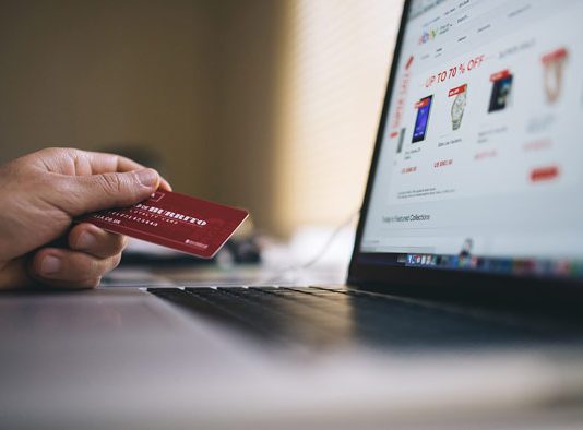 Una persona sostiene una tarjeta de crédito frente a una computadora portátil, navegando por un sitio web de compras en línea que muestra las últimas tendencias y descuentos exclusivos en comercio electrónico.