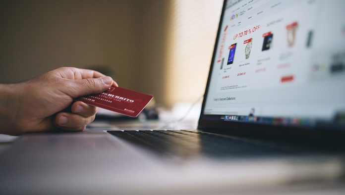 Una persona sostiene una tarjeta de crédito frente a una computadora portátil, navegando por un sitio web de compras en línea que muestra las últimas tendencias y descuentos exclusivos en comercio electrónico.