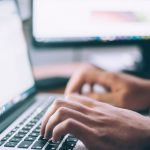 Una persona está escribiendo en un teclado portátil con un monitor de computadora en el fondo, tal vez navegando a través de interminables jornadas laborables.