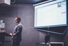 Una persona se encuentra de pie en un podio con una computadora portátil, presentando información web diseñada por expertos en una pantalla en una habitación poco iluminada.