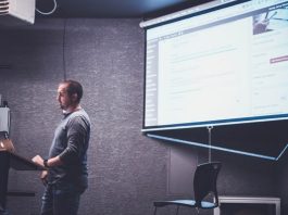 Una persona se encuentra de pie en un podio con una computadora portátil, presentando información web diseñada por expertos en una pantalla en una habitación poco iluminada.