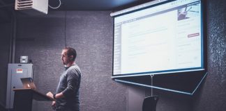 Una persona se encuentra de pie en un podio con una computadora portátil, presentando información web diseñada por expertos en una pantalla en una habitación poco iluminada.