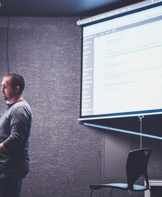 Una persona se encuentra de pie en un podio con una computadora portátil, presentando información web diseñada por expertos en una pantalla en una habitación poco iluminada.