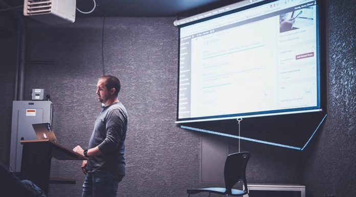 Una persona se encuentra de pie en un podio con una computadora portátil, presentando información web diseñada por expertos en una pantalla en una habitación poco iluminada.