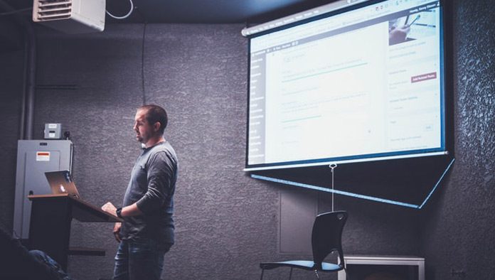 Una persona se encuentra de pie en un podio con una computadora portátil, presentando información web diseñada por expertos en una pantalla en una habitación poco iluminada.