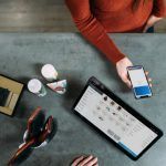 Una persona sostiene un teléfono inteligente sobre una mesa adornada con una tableta, cepillos, artículos de aseo y las máscaras esenciales que suelen utilizar los trabajadores esenciales.