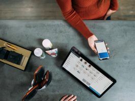 Una persona sostiene un teléfono inteligente sobre una mesa adornada con una tableta, cepillos, artículos de aseo y las máscaras esenciales que suelen utilizar los trabajadores esenciales.