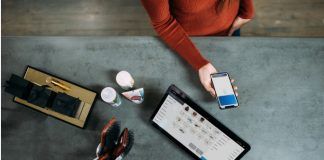 Una persona sostiene un teléfono inteligente sobre una mesa adornada con una tableta, cepillos, artículos de aseo y las máscaras esenciales que suelen utilizar los trabajadores esenciales.