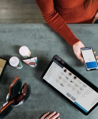 Una persona sostiene un teléfono inteligente sobre una mesa adornada con una tableta, cepillos, artículos de aseo y las máscaras esenciales que suelen utilizar los trabajadores esenciales.