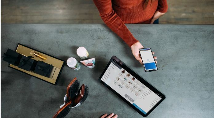 Una persona sostiene un teléfono inteligente sobre una mesa adornada con una tableta, cepillos, artículos de aseo y las máscaras esenciales que suelen utilizar los trabajadores esenciales.