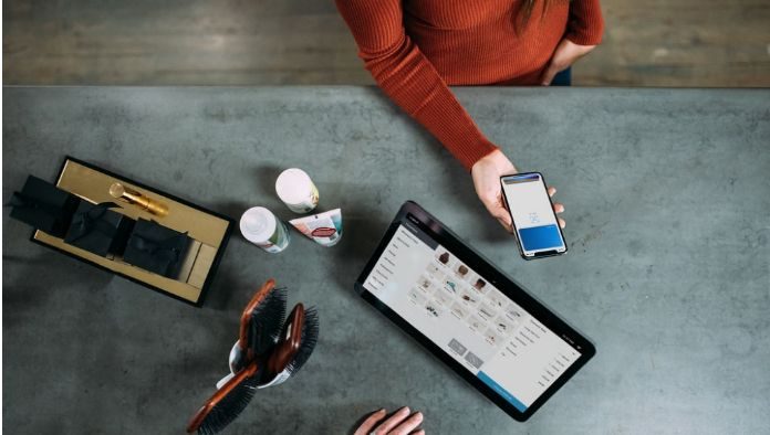 Una persona sostiene un teléfono inteligente sobre una mesa adornada con una tableta, cepillos, artículos de aseo y las máscaras esenciales que suelen utilizar los trabajadores esenciales.