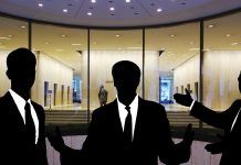 Siluetas de tres personas en traje discutiendo hábilmente sobre ventas técnicas frente a la entrada de un edificio con fachada de vidrio.