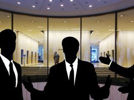 Siluetas de tres personas en traje discutiendo hábilmente sobre ventas técnicas frente a la entrada de un edificio con fachada de vidrio.