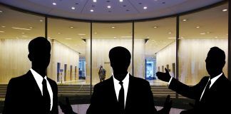 Siluetas de tres personas en traje discutiendo hábilmente sobre ventas técnicas frente a la entrada de un edificio con fachada de vidrio.