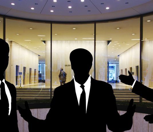 Siluetas de tres personas en traje discutiendo hábilmente sobre ventas técnicas frente a la entrada de un edificio con fachada de vidrio.
