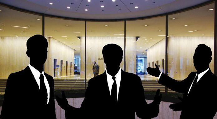 Siluetas de tres personas en traje discutiendo hábilmente sobre ventas técnicas frente a la entrada de un edificio con fachada de vidrio.