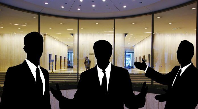 Siluetas de tres personas en traje discutiendo hábilmente sobre ventas técnicas frente a la entrada de un edificio con fachada de vidrio.