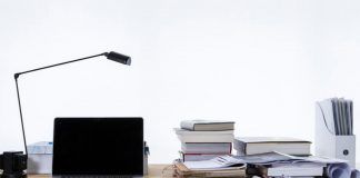 Dónde estudiar un Máster de Marketing Digital Un escritorio con una computadora portátil, una lámpara de escritorio y libros apilados sobre estrategias de marketing digital se encuentra junto a papeles y un archivador sobre un fondo liso.