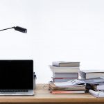 Un escritorio con una computadora portátil, una lámpara de escritorio y libros apilados sobre estrategias de marketing digital se encuentra junto a papeles y un archivador sobre un fondo liso.