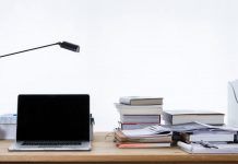 Un escritorio con una computadora portátil, una lámpara de escritorio y libros apilados sobre estrategias de marketing digital se encuentra junto a papeles y un archivador sobre un fondo liso.