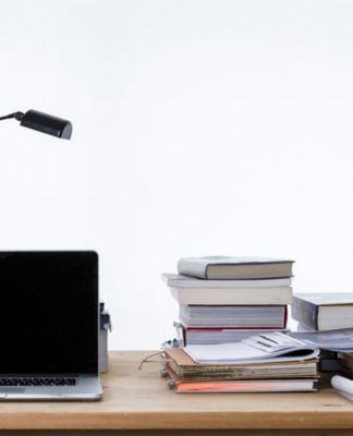 Un escritorio con una computadora portátil, una lámpara de escritorio y libros apilados sobre estrategias de marketing digital se encuentra junto a papeles y un archivador sobre un fondo liso.