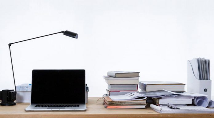 Dónde estudiar un Máster de Marketing Digital Un escritorio con una computadora portátil, una lámpara de escritorio y libros apilados sobre estrategias de marketing digital se encuentra junto a papeles y un archivador sobre un fondo liso.