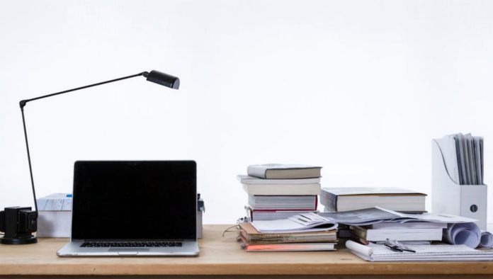 Diseño sin título Un escritorio con una computadora portátil, una lámpara de escritorio y libros apilados sobre estrategias de marketing digital se encuentra junto a papeles y un archivador sobre un fondo liso.