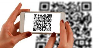 Una persona escanea un código QR con un teléfono inteligente, frente a un código QR más grande que aparece como fondo borroso, fusionando a la perfección los ámbitos de la interacción fuera de línea con las estrategias de marketing digital.