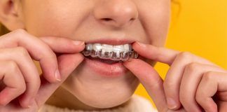 Un primer plano muestra a una persona colocando un alineador de ortodoncia transparente sobre sus dientes superiores sobre un fondo amarillo vivo, resaltando la precisión y el cuidado típicos de una clínica dental de renombre.