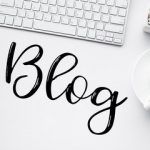 Un escritorio blanco contiene un teclado, un cactus, una taza de café y un bloc de notas. La palabra "Blog" está escrita con elegancia en letra cursiva grande, capturando la esencia de un espacio de trabajo con mucha tecnología listo para inspirar la creación de su última página web.