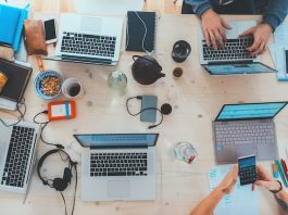 Vista aérea de un escritorio de trabajo lleno de computadoras portátiles, cuadernos, teléfonos, auriculares y bocadillos. Varias personas trabajan y usan varios dispositivos.