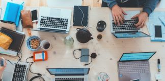 Vista aérea de un escritorio de trabajo lleno de computadoras portátiles, cuadernos, teléfonos, auriculares y bocadillos. Varias personas trabajan y usan varios dispositivos.