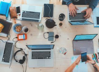 Vista a&eacute;rea de un escritorio de trabajo lleno de computadoras port&aacute;tiles, cuadernos, tel&eacute;fonos, auriculares y bocadillos. Varias personas trabajan y usan varios dispositivos.