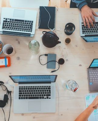 Vista aérea de un escritorio de trabajo lleno de computadoras portátiles, cuadernos, teléfonos, auriculares y bocadillos. Varias personas trabajan y usan varios dispositivos.