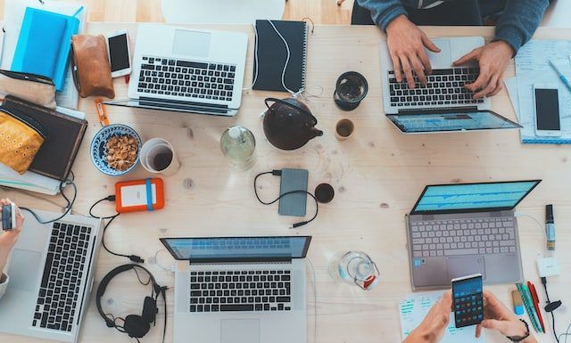 La importancia del diseño y el mantenimiento web Vista aérea de un escritorio de trabajo lleno de computadoras portátiles, cuadernos, teléfonos, auriculares y bocadillos. Varias personas trabajan y usan varios dispositivos.