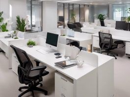 Espacio de oficina moderno con escritorios blancos, sillas negras, computadoras y plantas verdes. Mamparas de vidrio dividen áreas. Iluminación brillante y ambiente ordenado y limpio.