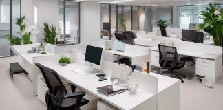 Gestoría online: Beneficios y consideraciones Espacio de oficina moderno con escritorios blancos, sillas negras, computadoras y plantas verdes. Mamparas de vidrio dividen áreas. Iluminación brillante y ambiente ordenado y limpio.