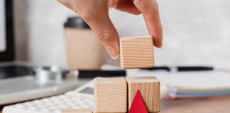 Externalizar la fuerza de ventas: una estrategia para crecer sin límites Una mano coloca un bloque de madera en una pila junto a una flecha roja, con una calculadora y una taza de café en el fondo.